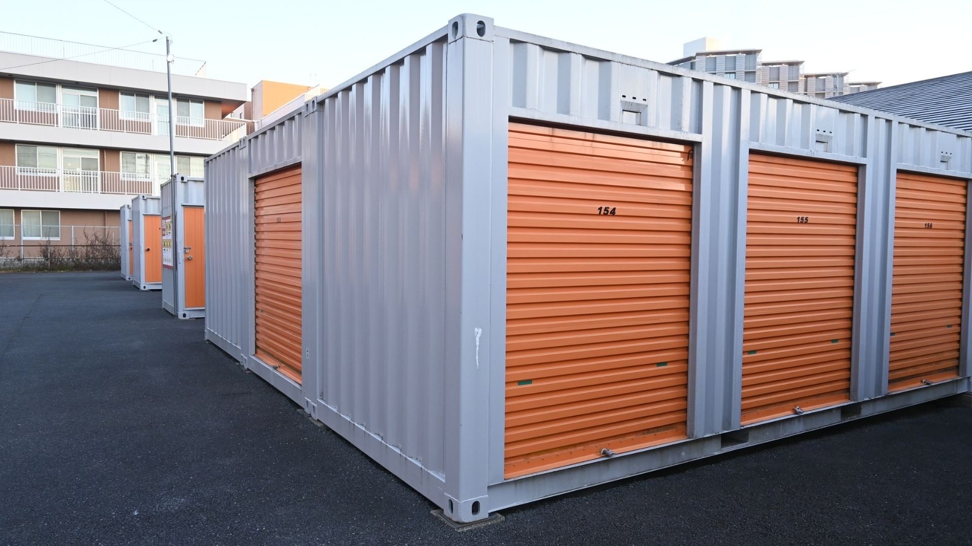 Row of metal storage units with orange roller doors and numbered compartments in urban setting