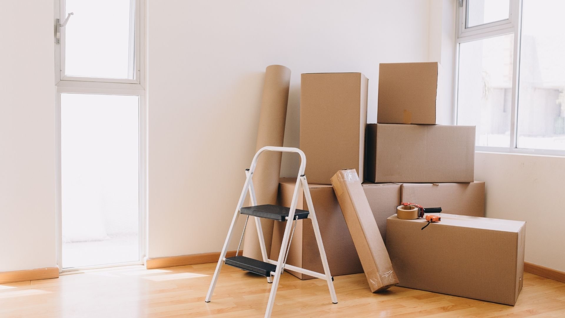 Bright empty room with cardboard moving boxes, step ladder, and packing supplies stacked together