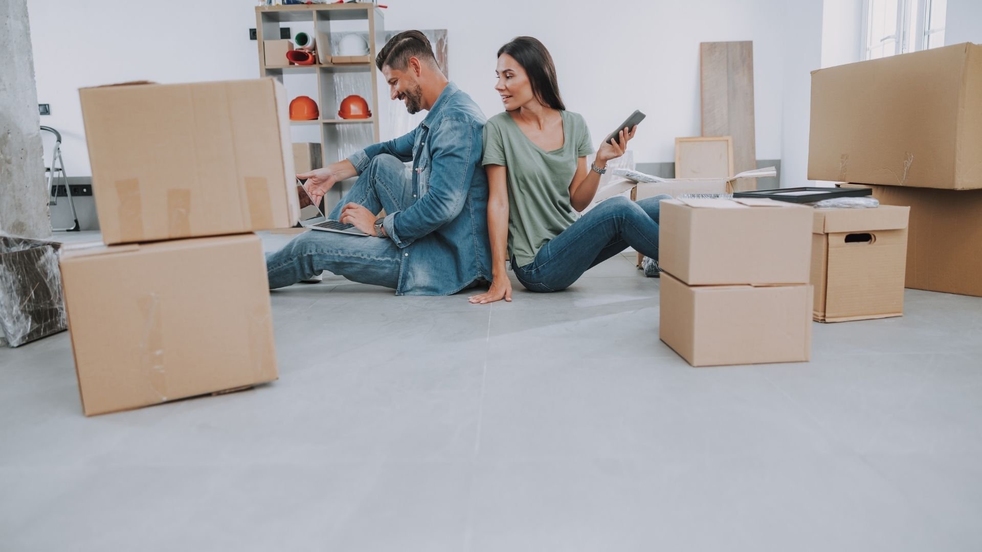 Couple sitting on floor surrounded by cardboard boxes, planning their move with tablet and smartphone.