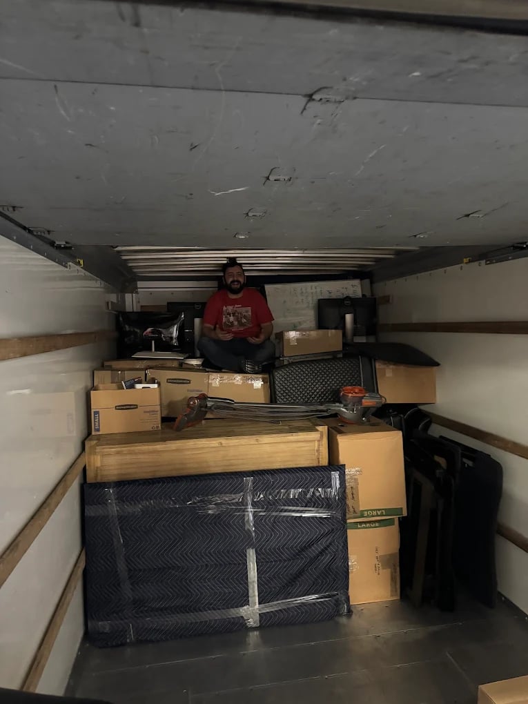 Man surrounded by cardboard boxes and furniture in crowded, cluttered moving truck interior